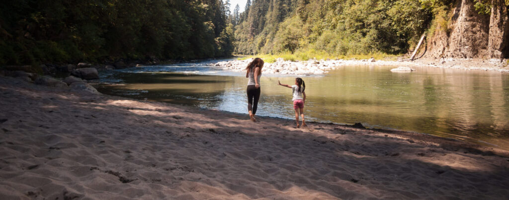 A mother and daughter are playing together along the beautiful banks of the Sandy River in Oregon. The little girl is playfully throwing sand at her mother, as she chases her.